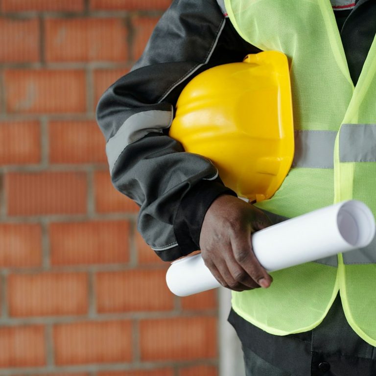Person wearing a yellow hard hat and reflective vest, holding construction plans.